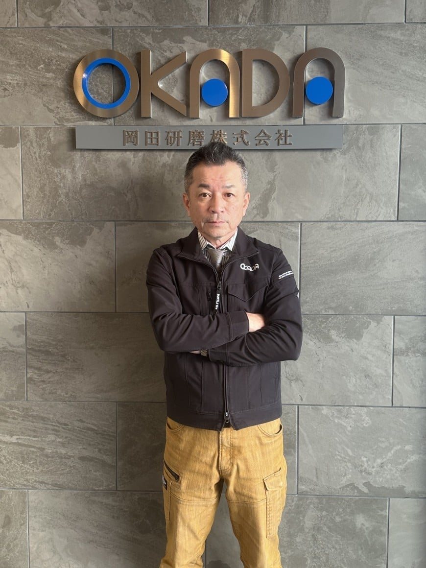 岡田研磨株式会社 代表取締役社長 岡田健一。OKADAのロゴ壁の前に立つ